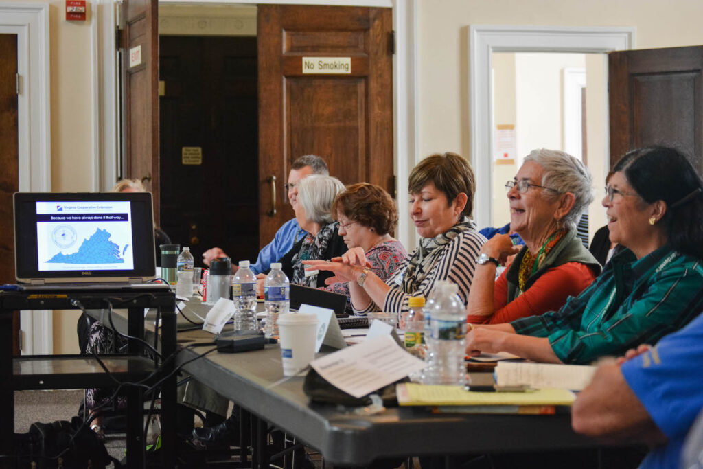 row of adults sit at a long table facing in front of them. Some laugh, some look down at papers, one checks a watch. An open laptop sits in front of the table displaying a screen with a map of Virginia.